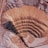 An aerial view of a large open-pit mine featuring concentric terraces carved into the earth, surrounded by barren, dusty terrain. There is heavy machinery at the bottom of the pit indicating active mining operations. The landscape is expansive and marked by shades of brown and pink.