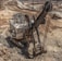 A large excavator is operating in a vast open-pit mining area, surrounded by dirt roads and rocky terrain. The machinery is actively digging into the earth, with dust rising from the operation. The landscape is vast and barren, showing multiple layers and pathways within the mine.